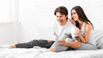A happy couple relaxes in bed, the man is holding a laptop while the woman holds a mug of coffee and smiles. The couple is enjoying their time together in a cozy setting, copy space photo