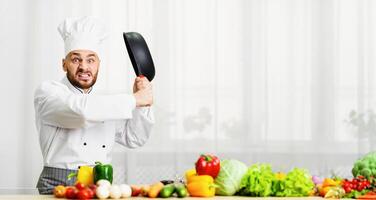 Mad Professional Chef Guy Raising Frying Pan Threateningly Grimacing Standing In Kitchen. Free Space For Text photo