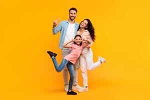 A happy family of three poses together in front of a yellow background. The father is on the left, making a peace sign with his hand, while the mother is on the right, with a bright smile. photo