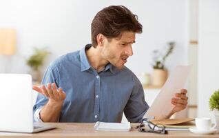 A man sits at a desk looking perplexed while reading a document. His expression suggests he is unhappy or surprised by the contents. photo