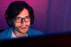 A man wearing headphones and glasses sits in a dimly lit room, concentrating intently on a computer screen in front of him. The soft, purple and blue lighting casts a calming glow on his face photo