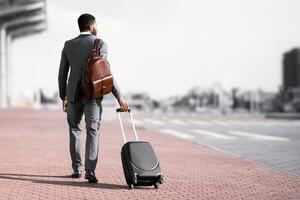 Back view of black businessman walking with rolling suitcase outside a modern building, ready for a business trip or travel. photo