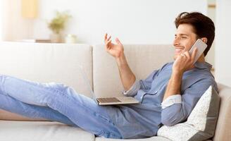 A man sits on a couch with a laptop in his lap and talks on a phone with a smile on his face. photo