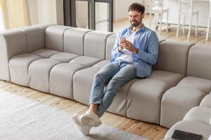 A man sits on a light grey modular couch in a living room, relaxed and smiling as he uses his phone. The man is dressed casually in a blue button-down shirt, white t-shirt, jeans, and white sneakers photo