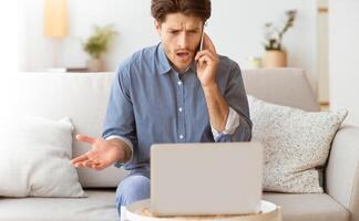 A frustrated man sits on a couch, looking at his laptop screen while talking on his phone. He is likely having technical difficulties. photo