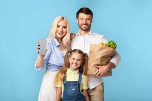 A happy family of three stands in front of a light blue background. The mother holds a smartphone in her right hand and smiles at the camera. The father holds a paper bag full of groceries photo