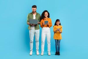 A man, woman, and young girl stand in a row against a light blue background. Family using various devices. Gadgets addiction photo