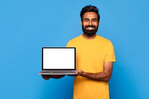 A bearded Indian man in a yellow t-shirt smiles while holding a laptop with a blank screen in front of him. He is standing in front of a solid blue background. photo