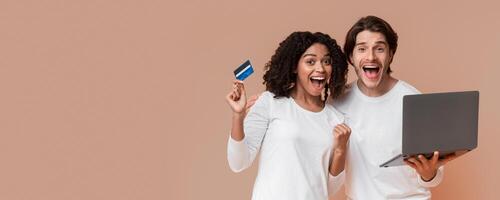 Online Shopping Concept. Overjoyed multiracial couple with laptop and credit card posing over yellow studio background, copy space photo