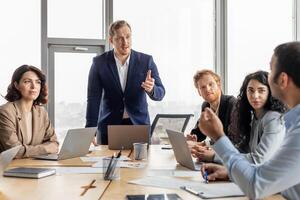 A group of business professionals are gathered around a conference table in a modern office. The presenter, standing at the head of the table, is speaking with passion and enthusiasm photo