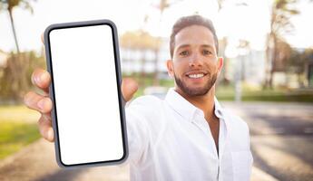 A smiling man in a white shirt holds a smartphone with a blank screen in front of him. The phone is in focus, while the man is blurred in the background. photo
