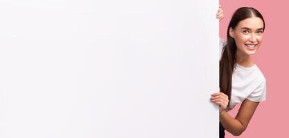 Smiling Millennial Girl Peeking Out Of Empty White Poster Posing In Studio Over Pink Background. Panorama, Mockup photo