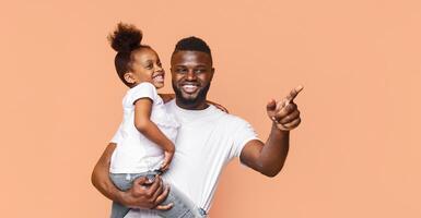 A happy Black father is holding his daughter in his arms while pointing to the right with a big smile on his face. The image captures a moment of joy and connection between a father and daughter. photo