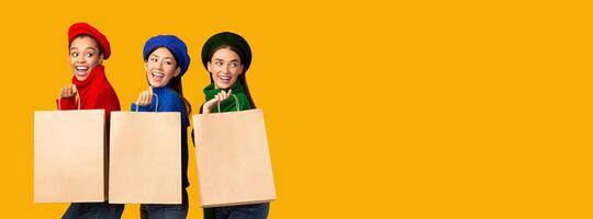 Three Happy Girls On Shopping Holding Paper Shopper Bags Standing Over Yellow Background In Studio. Mockup, Panorama photo