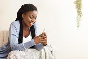 Smiling african american woman using smartphone at home, messaging or browsing social networks while relaxing on couch, copy space photo
