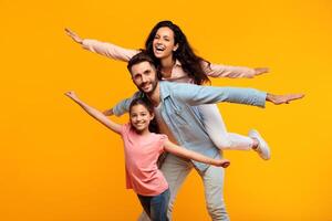 A happy family of three is playing airplane on a yellow background. The father is holding the mother on his back while the daughter is holding on to his shoulders. They are all smiling photo