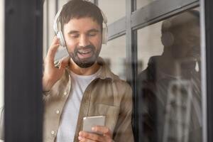 A man with a beard is singing along to music playing on his smartphone through headphones. He is standing near a window, and his reflection is visible in the glass. photo