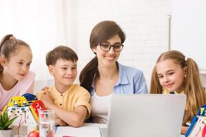 A teacher and three young students are gathered around a laptop computer in a classroom setting. They appear to be learning about robotics. The teacher is smiling and pointing at the screen photo