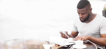 A man sits at a coffee shop table, looking at his phone while holding a notebook and pen. He appears to be reading something on his phone. photo