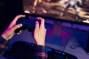 This is a photo of a persons hands holding a game controller, with a computer monitor in the background displaying an action game. The person is immersed in the game, focused on the screen.