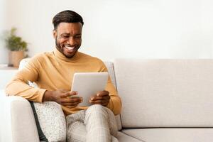 Great App. Smiling Afro Guy Using Tablet Computer Sitting On Couch At Home. Empty Space, Selective Focus photo