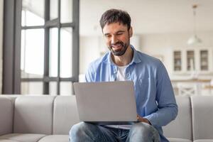 A man sits on couch in a modern home, smiling as he uses a laptop computer. He is dressed casually in a blue shirt and jeans. The home is bright and airy, with large windows letting in natural light. photo