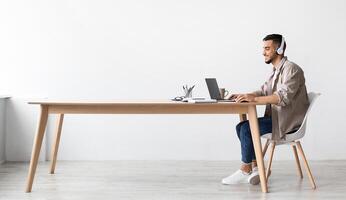 Cheerful Middle Eastern Freelancer Man Working On Pc Online At Home Listening To Music With Wireless Headphones, Making Research For New Project, Sitting At Desk Enjoying Remote Job, Profile Side View photo