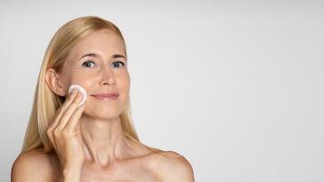 A blonde woman with bare shoulders is shown applying a cotton pad to her cheek. She has a light complexion and appears to be in her 40s. She has a gentle smile on her face, copy space photo