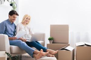 A young couple sits on a couch in their new home, surrounded by boxes. They are looking at a laptop and smiling. photo