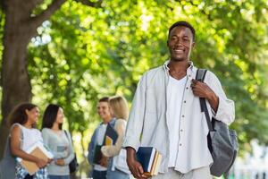 Happy black student with books smiling at camera, spending time with his friends in public park, copy space photo