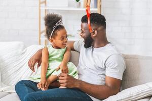 A father and daughter are sitting on a couch together. The daughter is wearing a princess crown and a green dress. The father is wearing a white T-shirt. Both are looking at each other and smiling. photo