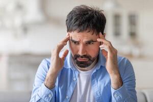 The image shows a man with a beard and short dark hair, wearing a light blue button-down shirt, sitting in a home setting with his hands on his temples. He has a concerned expression, closeup photo
