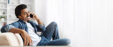 African American man enjoys a moment of relaxation on the couch, engaged in a cheerful phone conversation at his home during the afternoon, surrounded by soft light and a cozy atmosphere. photo