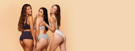 Three multiracial young women in stylish activewear smiling and posing playfully, exuding confidence and camaraderie, has fun on beige backdrop. Confidence, wellness, friendship, copy space photo