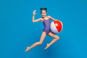 A young girl in a colorful bathing suit is standing on the beach, joyfully holding a beach ball. The vibrant beach ball contrasts with the blue sky and sandy shore. photo