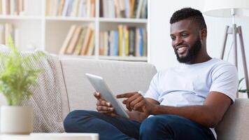 A smiling black man sits on a white couch in his living room. He is casually dressed and using a tablet computer, looking relaxed and enjoying his leisure time. The room is well-lit and decorated photo