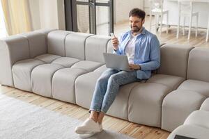 A man sits on a modern gray sectional sofa in a living room. He is looking at a smartphone and smiling, while using a laptop. He is wearing jeans and a blue button-up shirt, high angle view photo