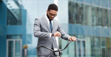 African American businessman on a bike in the city is checking his watch. The setting is urban and busy, capturing the essence of multitasking and staying on schedule in a fast-paced environment. photo