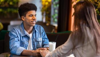 Black guy and girl are engaged in a conversation at a table in a restaurant. The guy appears to be speaking while the girl listens attentively. The setting suggests a casual dining environment. photo