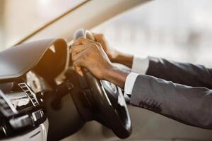 This image shows a closeup of a persons hands gripping the steering wheel of a car. The driver is wearing a suit jacket and their hands are visible in the foreground photo