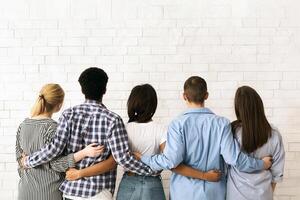 A group of multiethnic teenagers stands close together with their backs to the viewer, arms around each other in a display of friendship and unity, back view photo