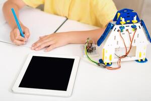 A child is sitting at a desk, learning about robotics and coding. They are using a tablet computer and a robot kit. The childs hand is shown writing on a piece of paper photo