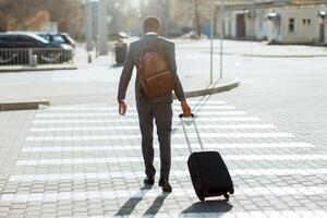 Black businessman in a suit crossing the street with a rolling suitcase, seen from behind in an urban setting. photo