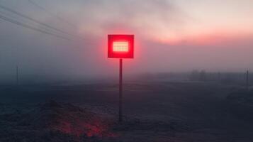 A red light, emanating from a square sign on a pole, illuminates the foggy landscape at sunrise. The sky is a soft pink, while the foreground is obscured by mist. photo