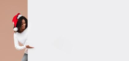 Cheerful Interracial Couple In Santa Hats Pointing At White Billboard With Free Space For Advertisement Or Text, Yellow Background photo