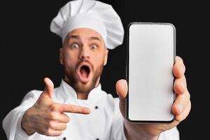 Culinary App. Excited Male Chef Showing Phone Screen Pointing Finger Recommending Application Posing On Black Studio Background. Mockup, Selective Focus photo