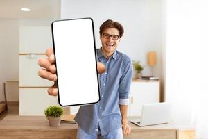 A man in a blue shirt holds a smartphone with a blank screen in front of him, smiling at the camera. He is standing in an office setting. photo