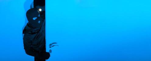 A cloaked individual cautiously opens a blue door in a darkened environment, illuminating the space with a flashlight, creating a suspenseful atmosphere of intrigue. photo