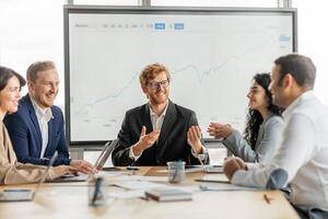 A group of diverse business professionals are gathered around a conference table in a modern office setting. They are looking at large screen displaying a growth chart, engaging in lively discussion photo