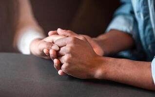 Two individuals are seated at a table, holding hands. Their interlocked fingers convey a sense of connection and intimacy. The table is simple, adding to the focus on the human interaction. photo